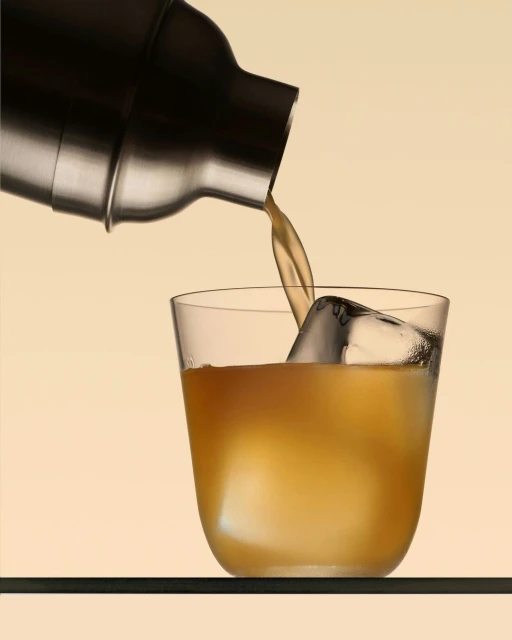 Drink being poured from a metal cocktail shaker into a clear glass with ice against a gradient light-yellow background. 