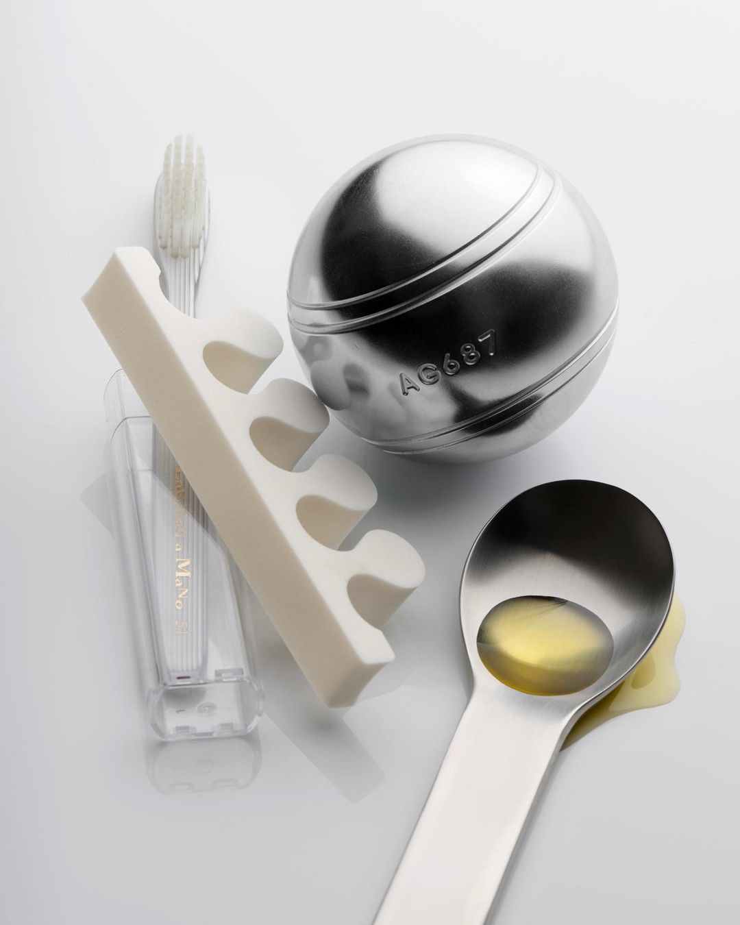 Still life of a metal ball, toe separators, toothbrush, and spoon filled with liquid against a white backdrop.