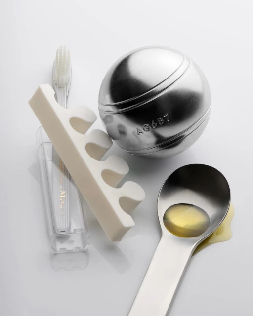 Still life of a metal ball, toe separators, toothbrush, and spoon filled with liquid against a white backdrop.
