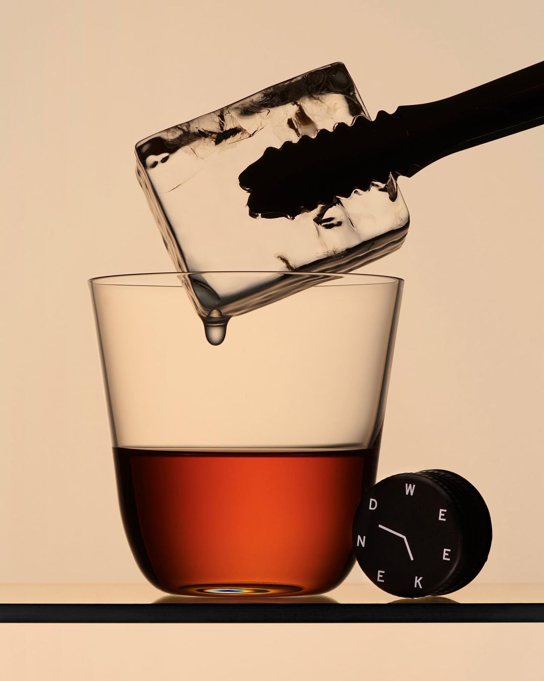 A clear glass of amber liquid with an ice cube held by black tongs, next to a black and white circular object against a neutral background.