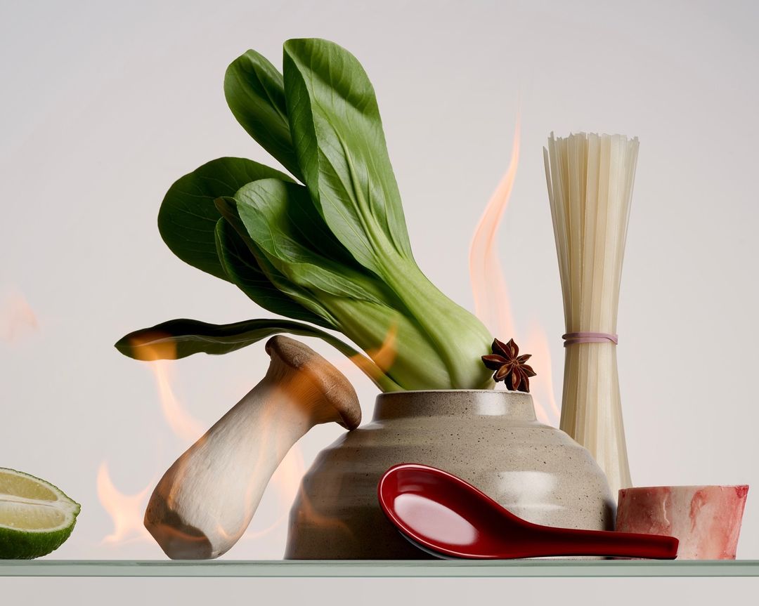 Stylized culinary still life features bok choy, mushroom, noodles, lime, bone, and red spoon in a pot, set against a white background with fire.