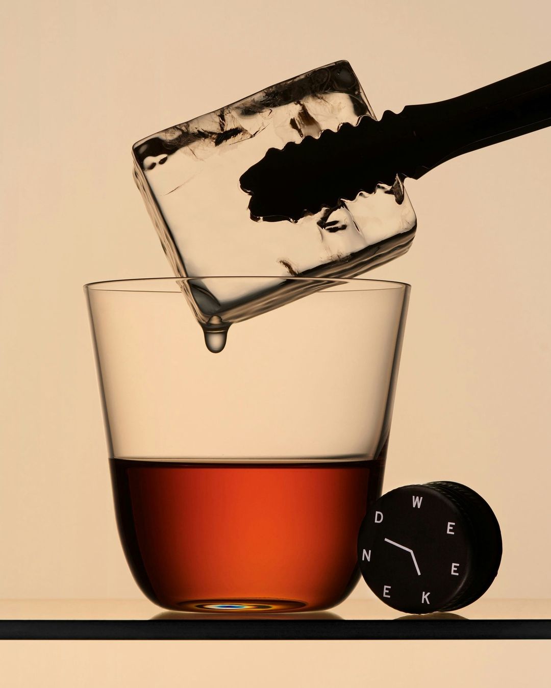 Still life shot of ice being dropped into a glass with amber liquid using ice tongs, accompanied by a clock on a light orange background.