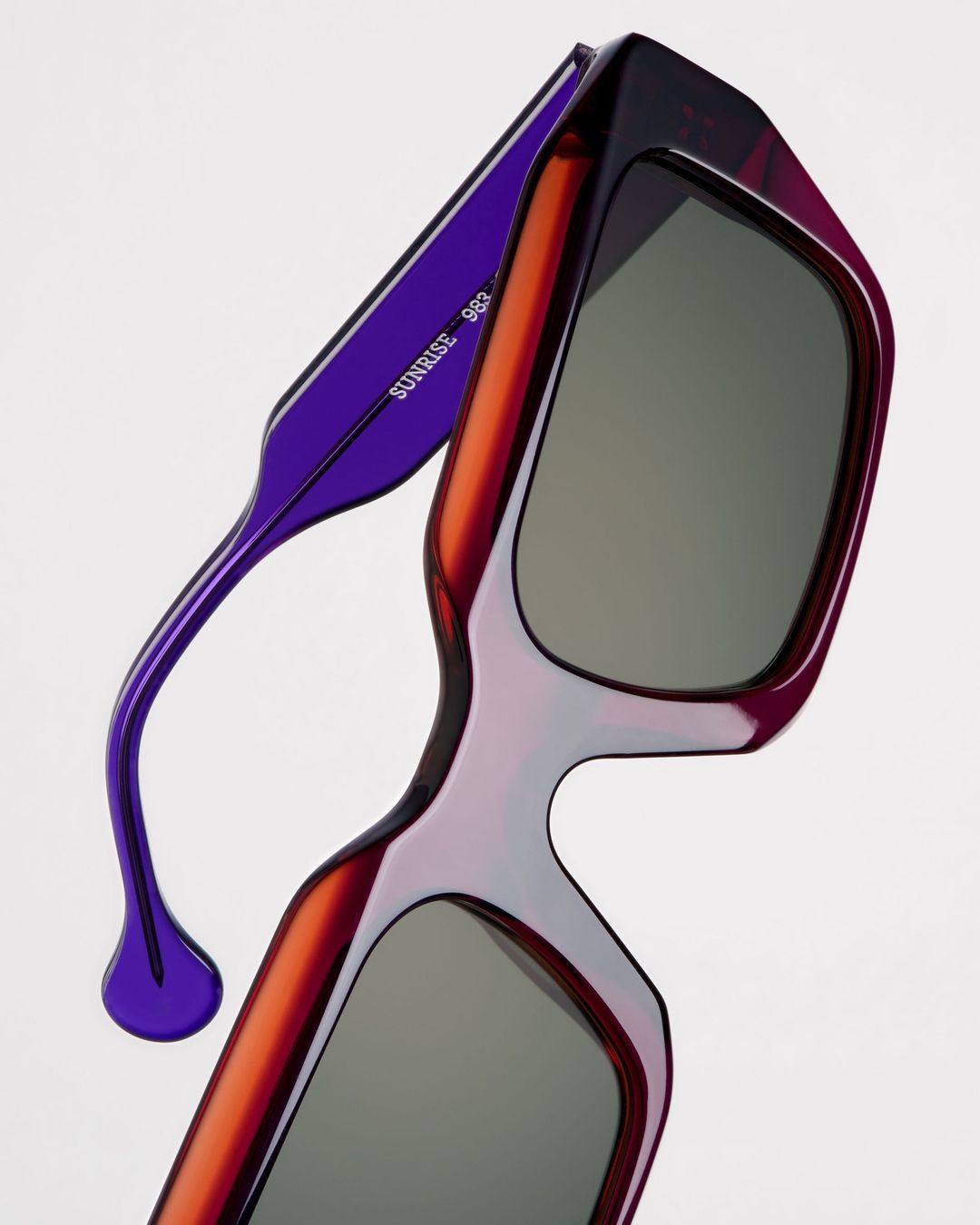 Close-up of purple and rust-colored sunglasses with gray lenses. Modern eyewear design against a pale backdrop.