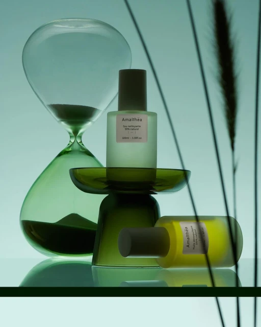 Still life studio shot with green-tinted hourglass and two cosmetic bottles on a reflective surface, in a minimalist beauty product presentation.