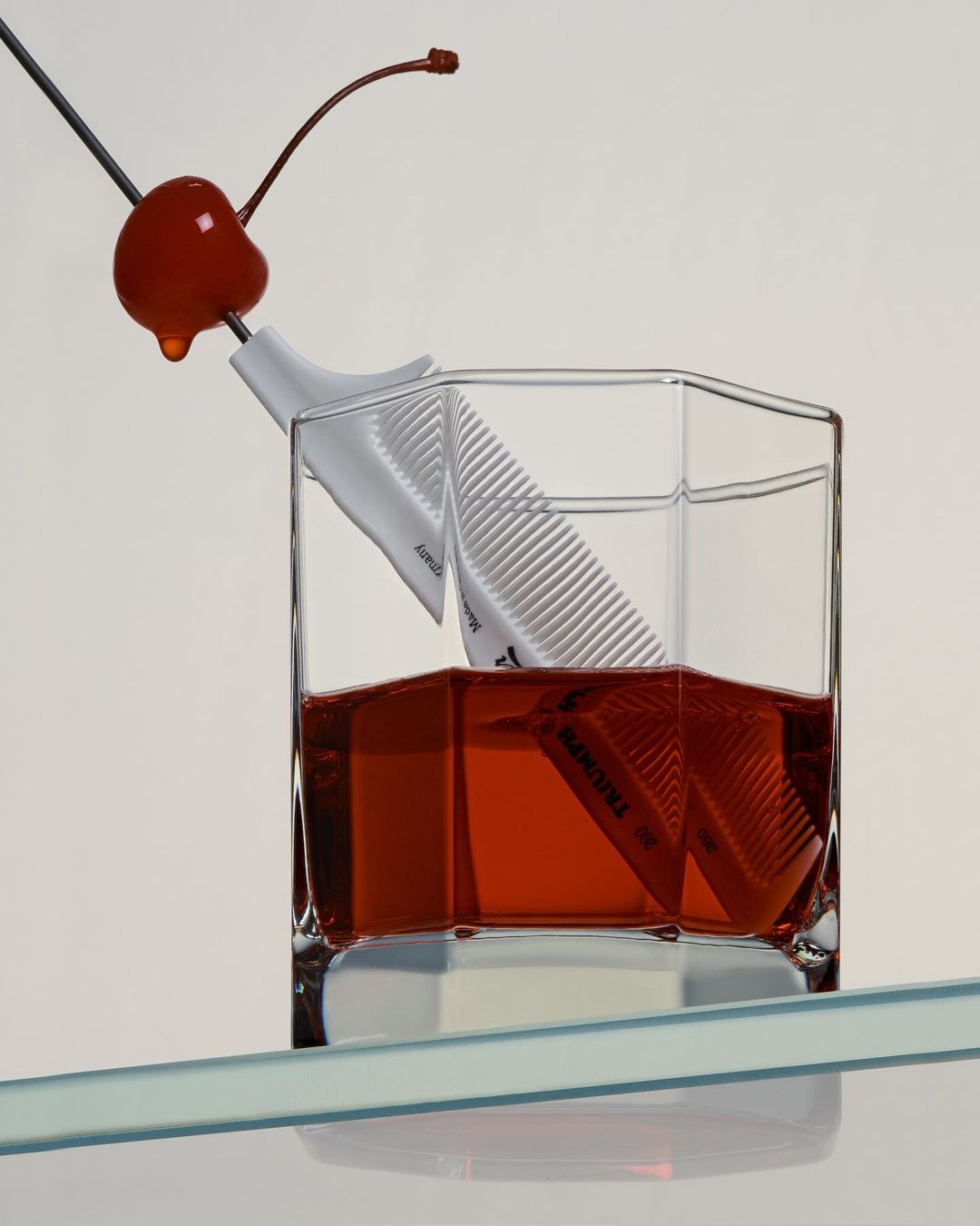 Still life of a cocktail glass filled with red liquid and two white combs, topped with a red cherry on a white surface.