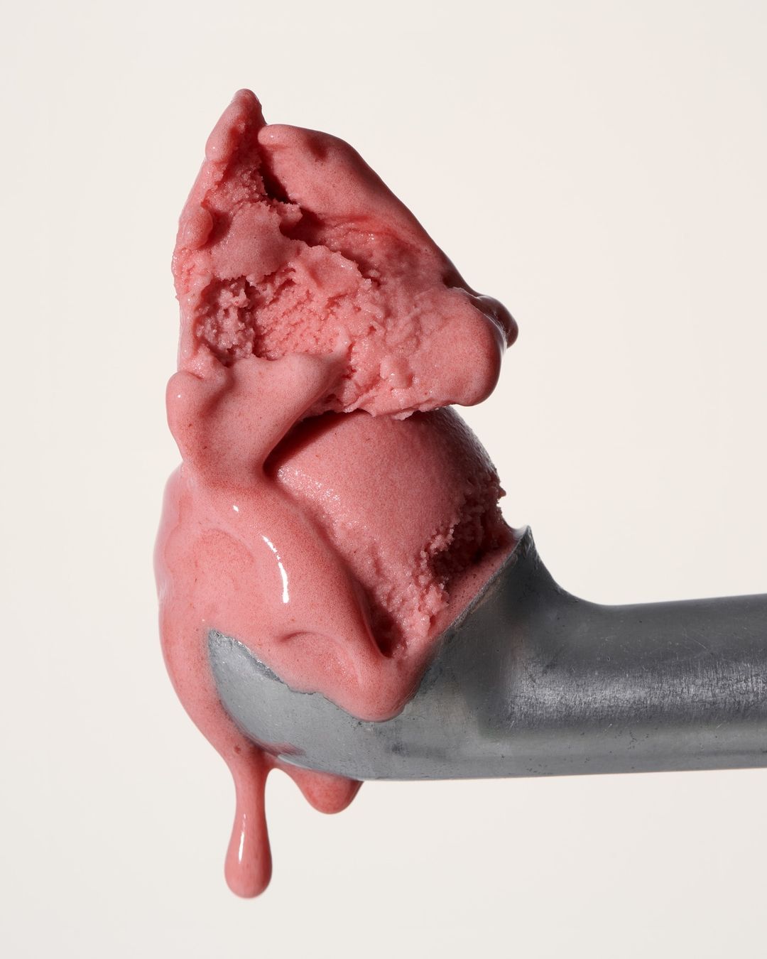 Close-up of a scoop of vibrant pink ice cream melting slightly, held by a silver ice cream scoop against a neutral background.