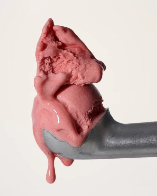 Close-up of a scoop of vibrant pink ice cream melting slightly, held by a silver ice cream scoop against a neutral background.