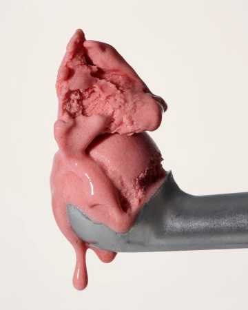 Close-up of a scoop of vibrant pink ice cream melting slightly, held by a silver ice cream scoop against a neutral background.