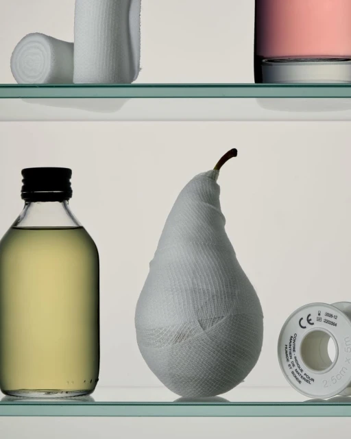 Still life of a pear wrapped in white bandage, on a glass shelf, with a small bottle and bandages in the background.