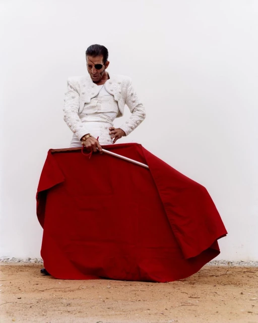 Matador poses with red cape in arena. Man with eyepatch wears traditional white suit against a plain background.