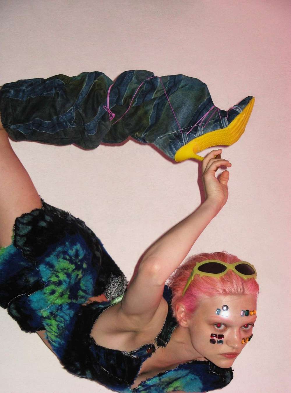 Woman in futuristic fashion wearing blue tie-dye outfit and tall yellow-soled boots posing in a photo studio against a pink background.