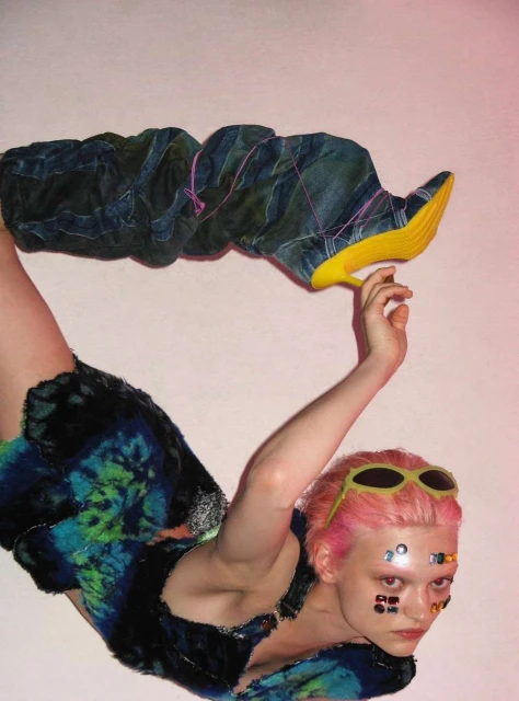Woman in futuristic fashion wearing blue tie-dye outfit and tall yellow-soled boots posing in a photo studio against a pink background.