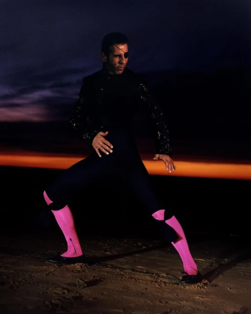 An adult male dancer in a black and pink outfit striking a pose on a beach at twilight with a moody sky in the background.