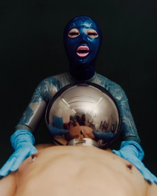 Surreal scene featuring a person in a blue latex mask and suit holding a reflective sphere over another person lying on their back.