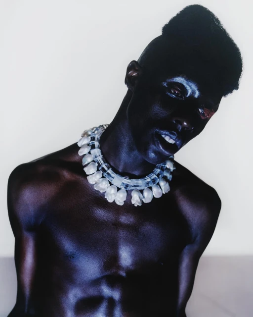 Close-up studio portrait of a Black man with dark skin, makeup, and a translucent beaded necklace against a bright, neutral backdrop.
