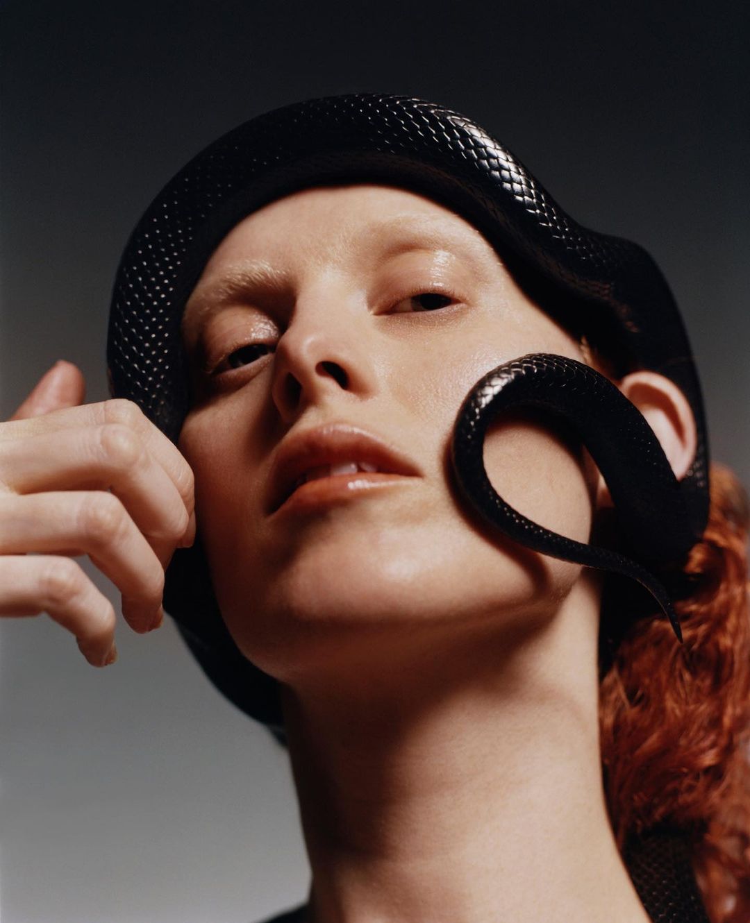 Portrait of a red-haired model wearing a snake as headwear, set against a grey backdrop. The snake's scales are visible.