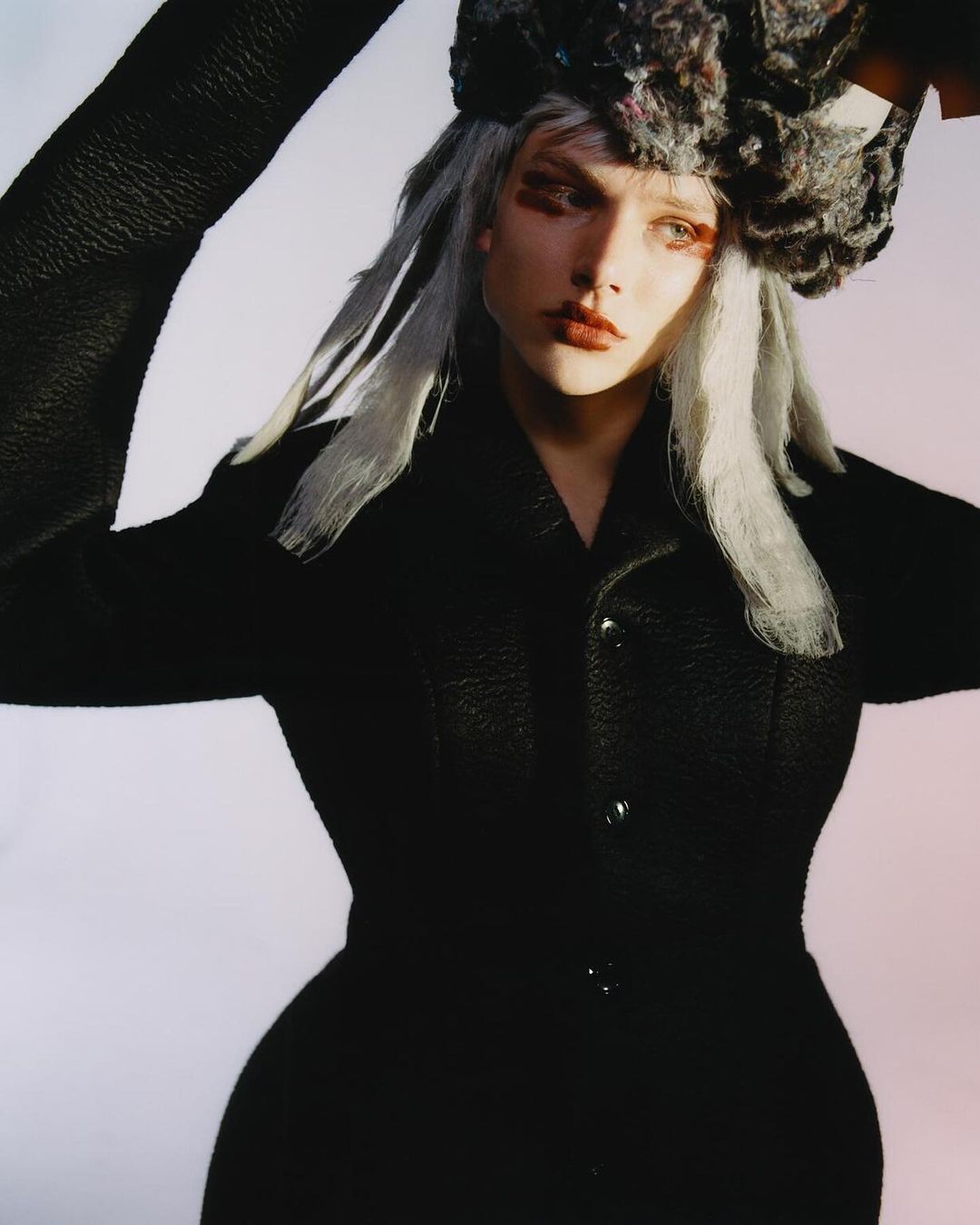 A fashion-forward young adult woman poses, wearing a black textured jacket, an elaborate gray hat, and dramatic eye makeup against a pale background.