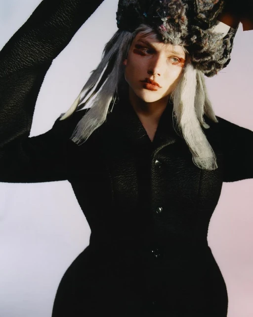 A fashion-forward young adult woman poses, wearing a black textured jacket, an elaborate gray hat, and dramatic eye makeup against a pale background.