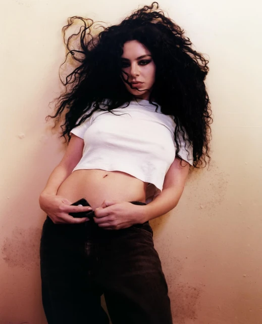 Brunette woman in a white top and dark jeans poses indoors. She has long, dark, wavy hair and smoky eye makeup.