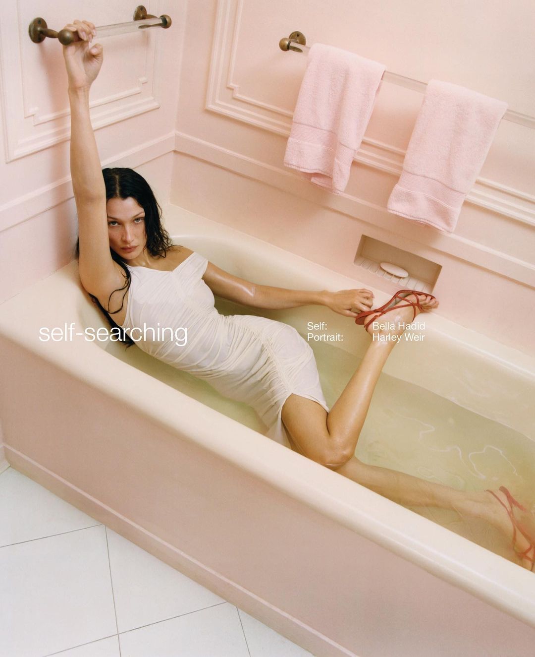 Woman in white dress poses in bathtub with red heels, holding onto a towel rack in a pink bathroom, bathed in soft light.