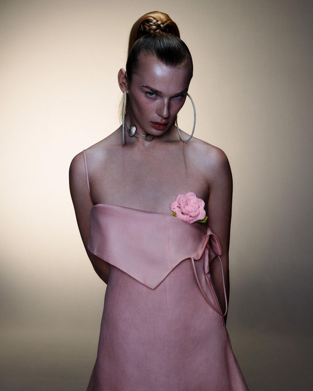 Fashion portrait of a woman with a braided ponytail wearing a pink strapless dress and statement jewelry in a neutral-toned studio.