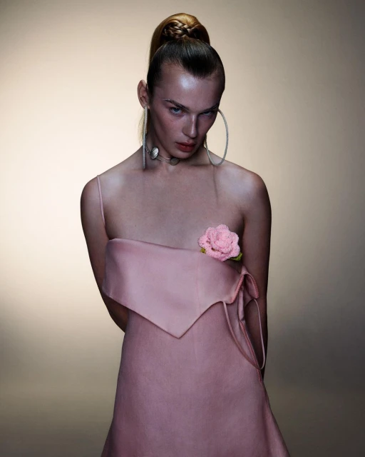 Fashion portrait of a woman with a braided ponytail wearing a pink strapless dress and statement jewelry in a neutral-toned studio.