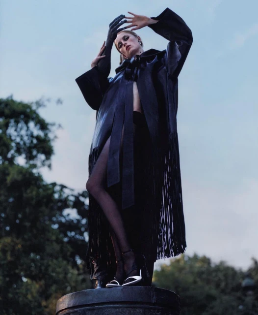 Adult female fashion model poses in an outdoor setting wearing a black coat with fringe and high heels, hands raised overhead.