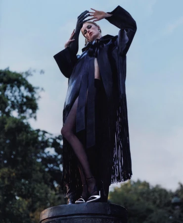 Adult female fashion model poses in an outdoor setting wearing a black coat with fringe and high heels, hands raised overhead.