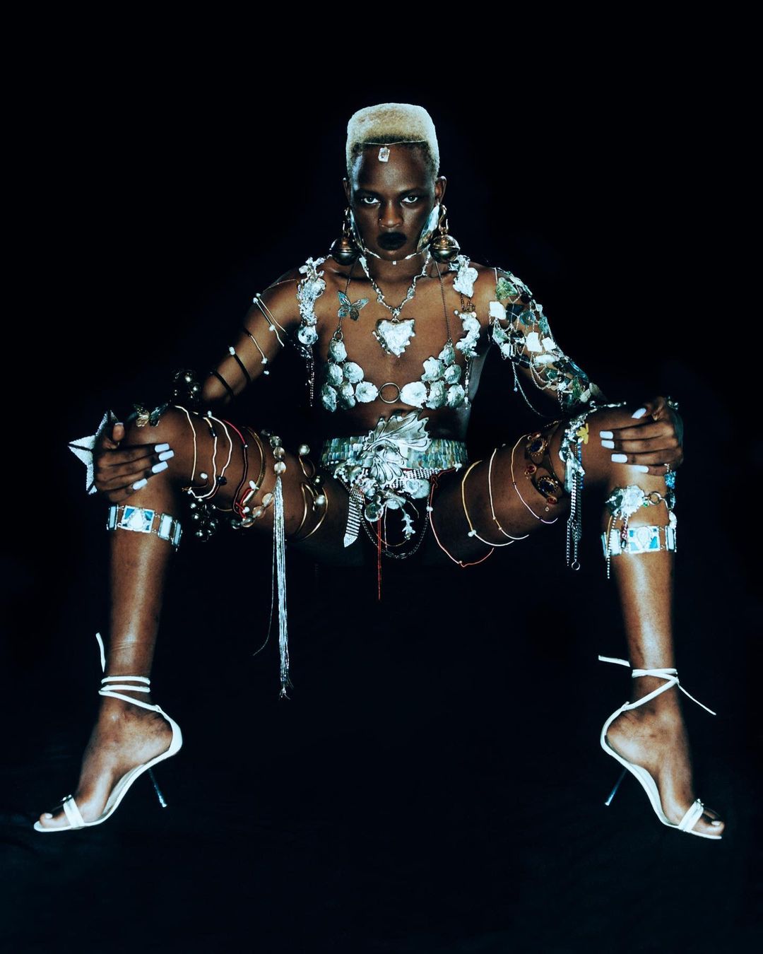 Elegant portrait of a Black woman with an unusual haircut, fashionable jewelry, and a nude bodysuit against a dark backdrop.