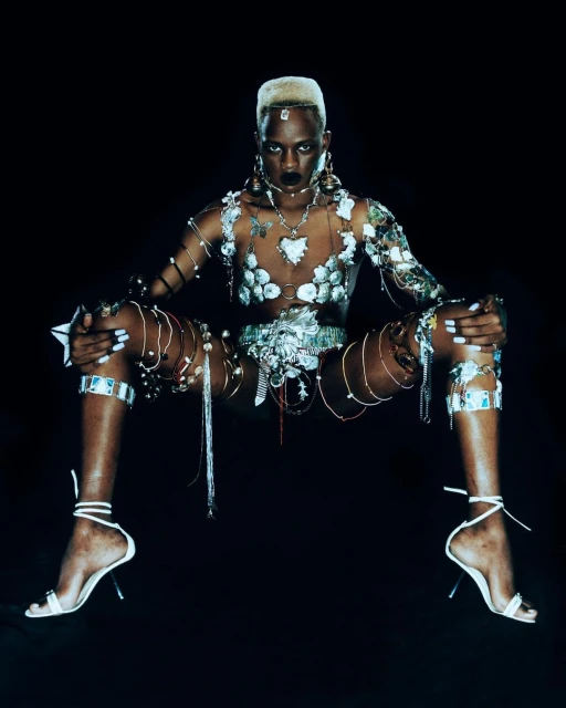 Elegant portrait of a Black woman with an unusual haircut, fashionable jewelry, and a nude bodysuit against a dark backdrop.