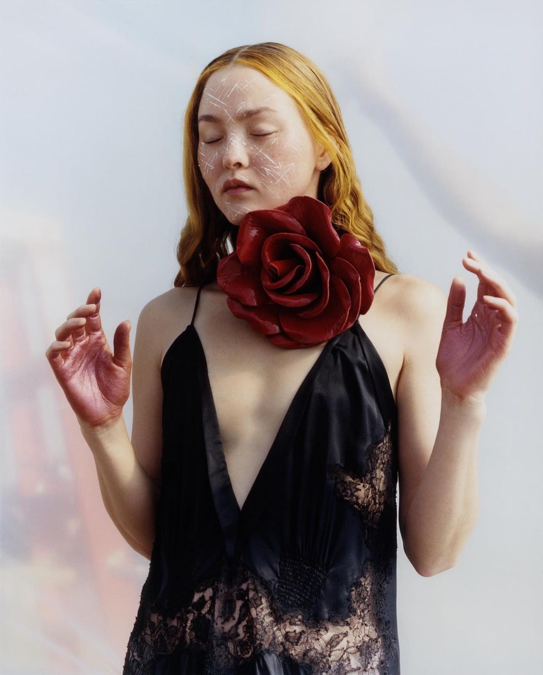 Woman with light skin and red hair, eyes closed, wearing a black dress and large red rose collar, hands raised, in a photo studio.