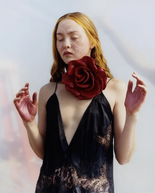 Woman with light skin and red hair, eyes closed, wearing a black dress and large red rose collar, hands raised, in a photo studio.