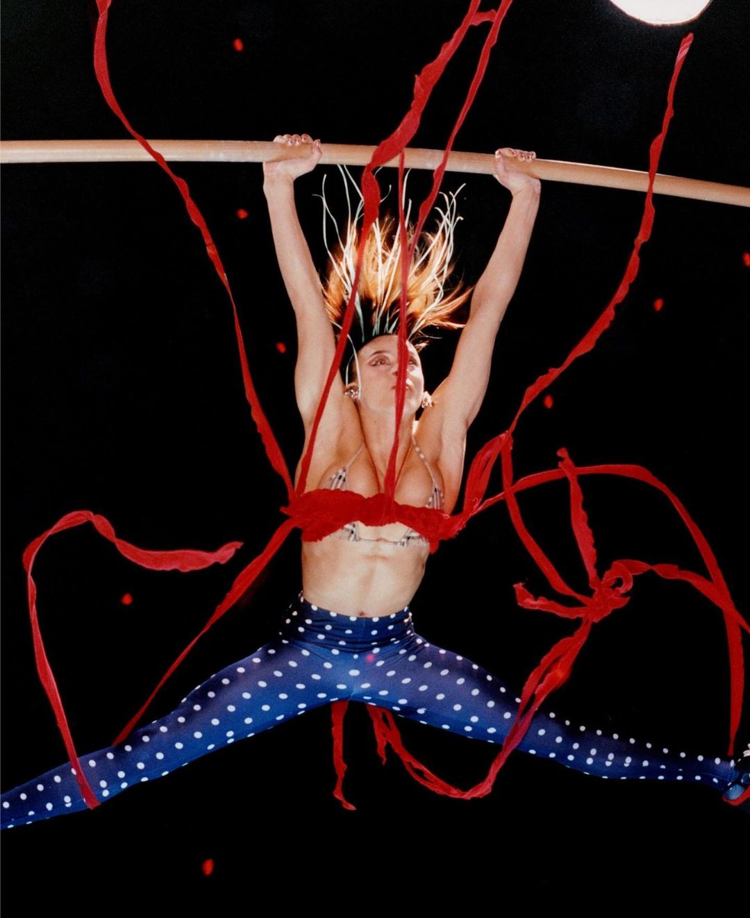 A woman in blue polka dot pants is hanging on a bar against a black background with red ribbon-like elements.