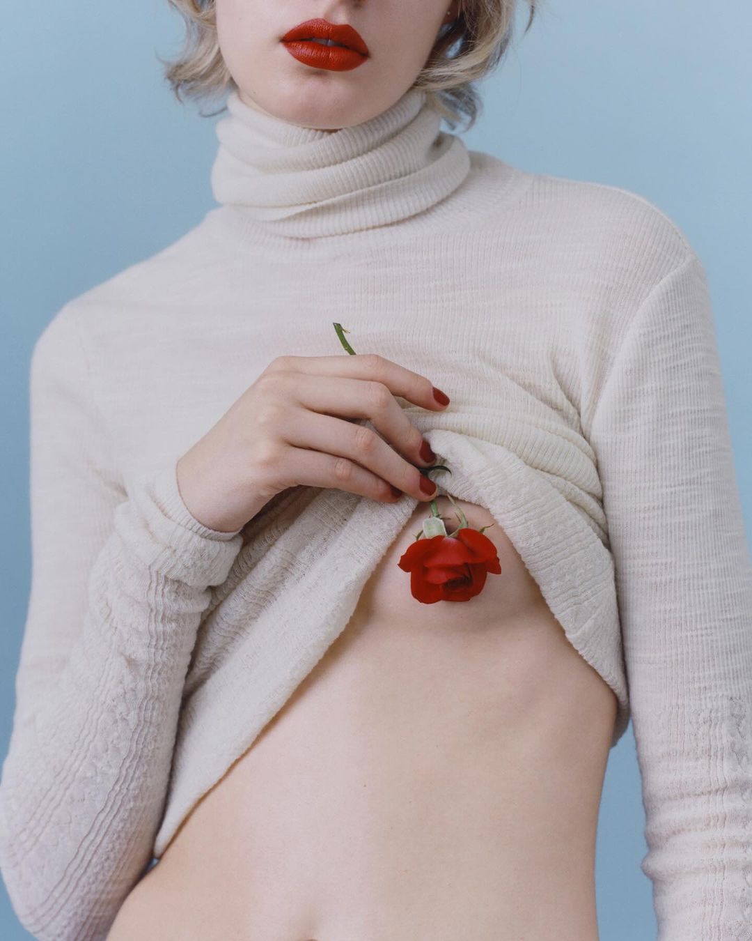 Torso shot of a young fair-skinned woman in a turtleneck sweater, pale lips, and a red rose over her chest on blue backdrop.