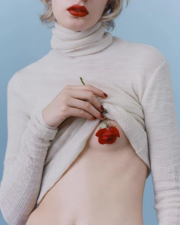 Torso shot of a young fair-skinned woman in a turtleneck sweater, pale lips, and a red rose over her chest on blue backdrop.