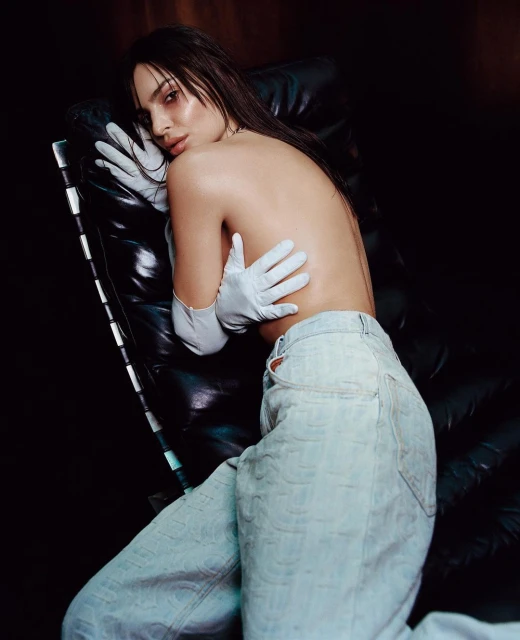 A woman with wet brunette hair, wearing jeans and white gloves, is reclining on a modern leather chair against a dark background.