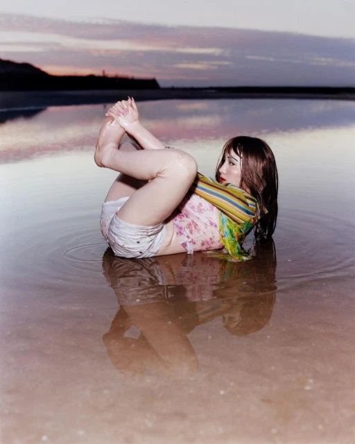 A woman sits in shallow water near the shore, holding her feet, with an orange and purple sunset in the background.