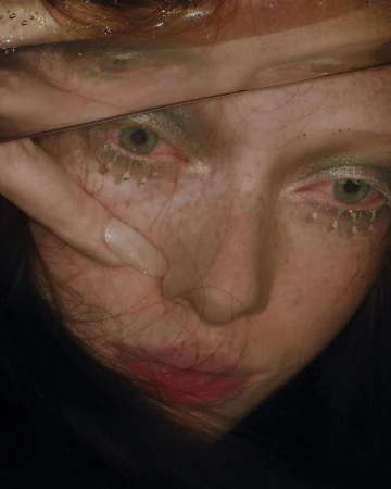 Close-up portrait of a woman with red hair, freckles, and green eye makeup viewed through a reflective surface by Harley Weir