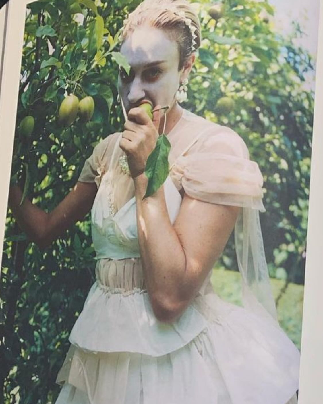 Woman in a light dress standing by a fruit tree in daylight, eating a green fruit. Outdoor setting with natural light and a dreamy aesthetic.