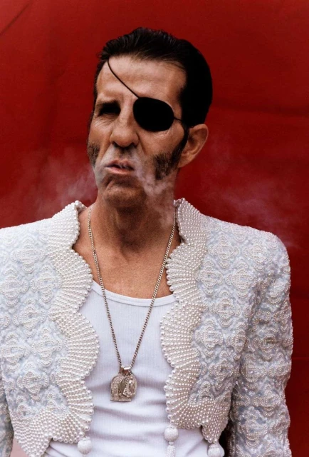 Adult male in matador costume with eyepatch smoking against a red background. Man wearing ornate white jacket with necklace.
