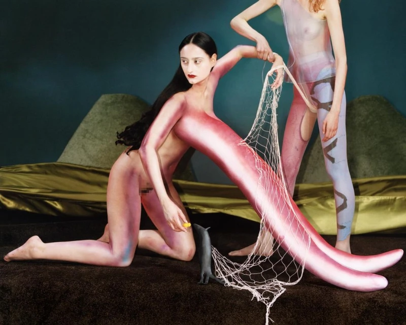 Two figures in a studio setting, one kneeling with pink body paint and one standing, holding a net; surreal, artistic, and theatrical.