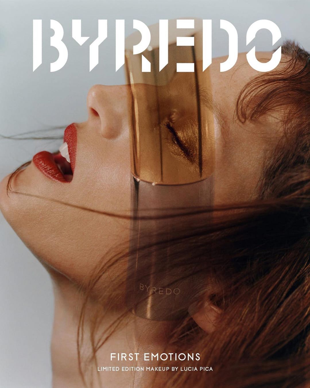 Fashion editorial featuring a model with red lips and a transparent gold face shield, alongside a BYREDO fragrance bottle.