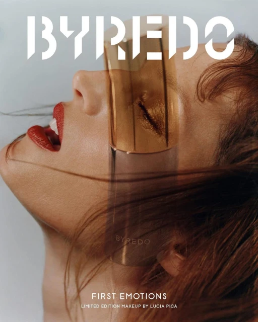 Fashion editorial featuring a model with red lips and a transparent gold face shield, alongside a BYREDO fragrance bottle.