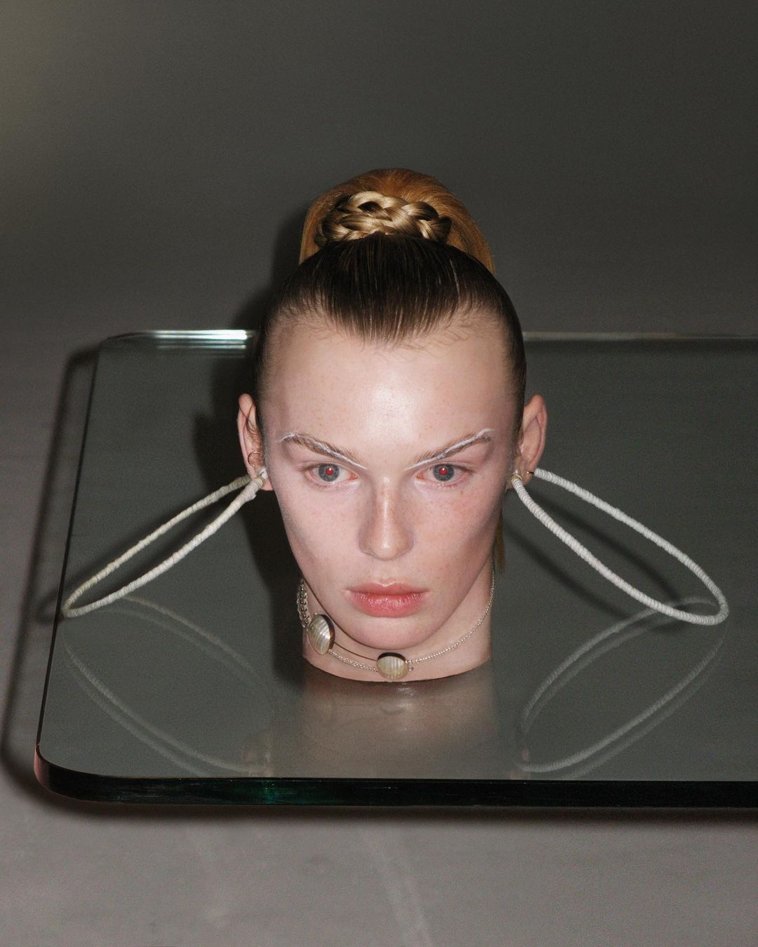 Conceptual fashion shot of a blonde female model with dramatic makeup, a modern necklace, and a futuristic hair style posing indoors.