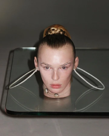 Conceptual fashion shot of a blonde female model with dramatic makeup, a modern necklace, and a futuristic hair style posing indoors.