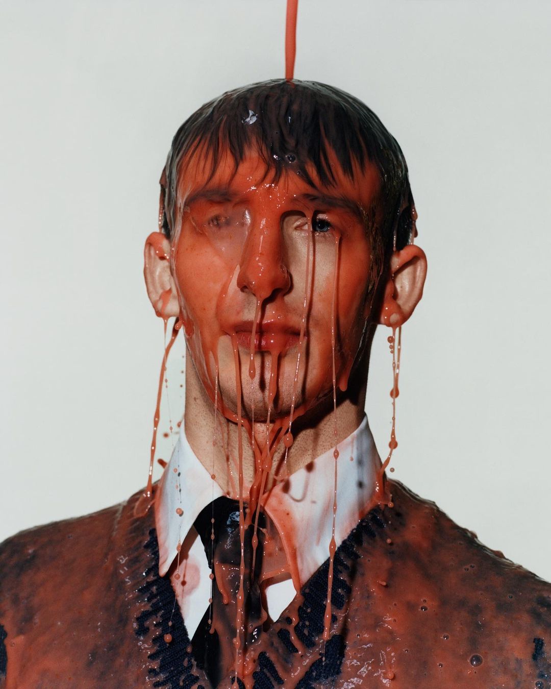 Portrait of an adult male with a neutral expression, covered in red liquid, wearing a white collared shirt and a dark v-neck sweater.