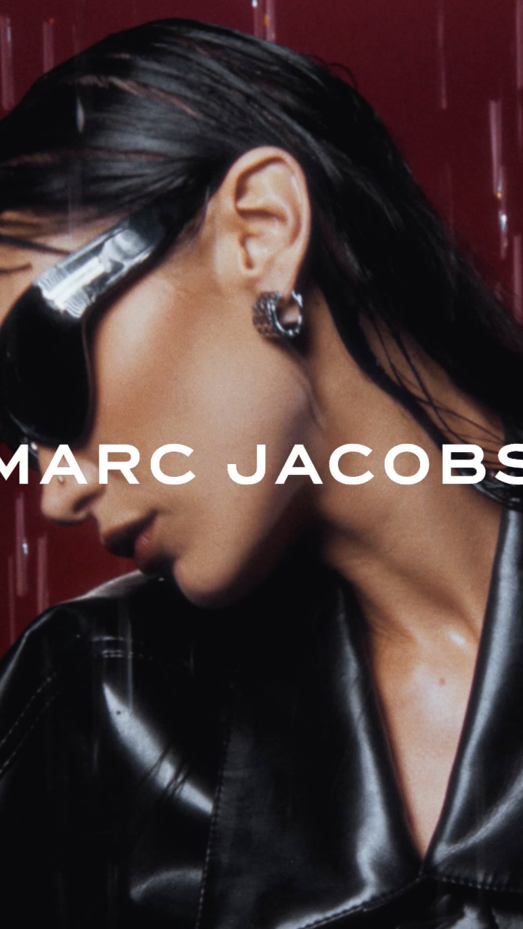 Close-up fashion shot of a woman with slicked-back black hair, wearing sunglasses and a black coat, with a ‘Marc Jacobs’ logo overlay.