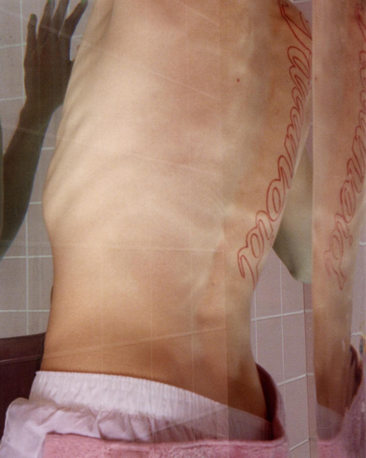Nude torso of an adult with a tattoo, reflected in glass with white tiled background and pink towel. Close-up, indoor setting.