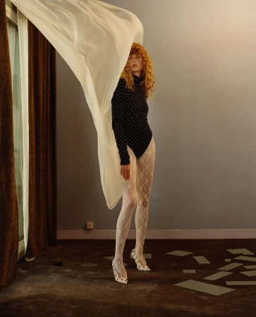 Fashion shot of a red-haired model in a black polka dot bodysuit and white patterned tights, with a flowing white curtain indoors.