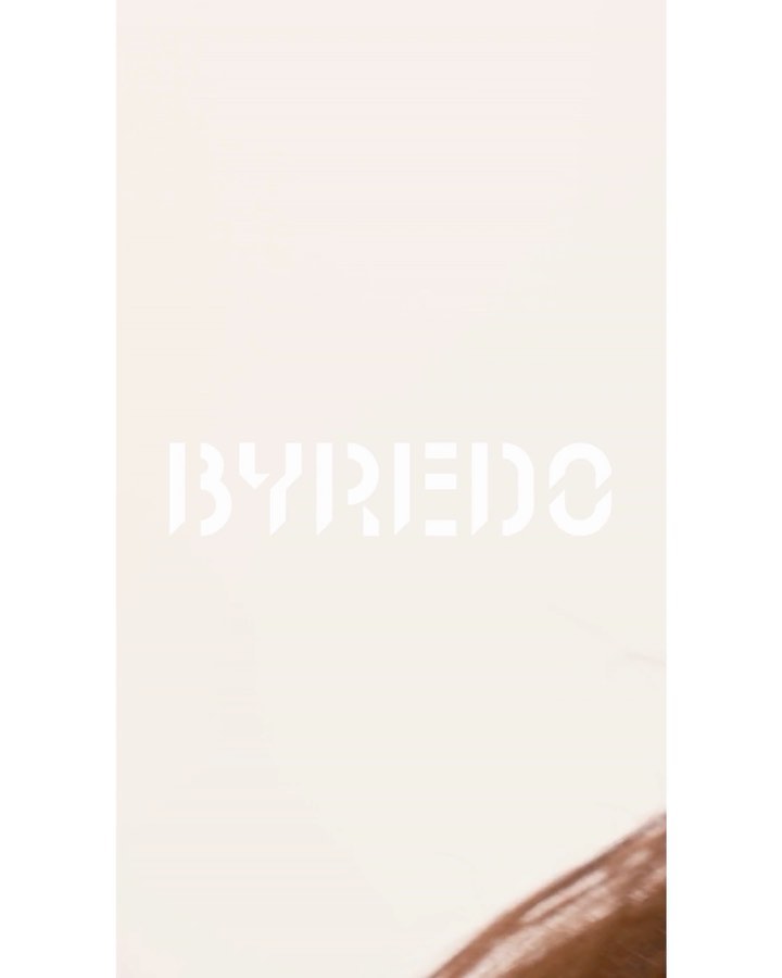 Close-up studio shot of light brown hair with dark roots, styled with a slight wave, against a beige background with the 'BYREDO' logo.
